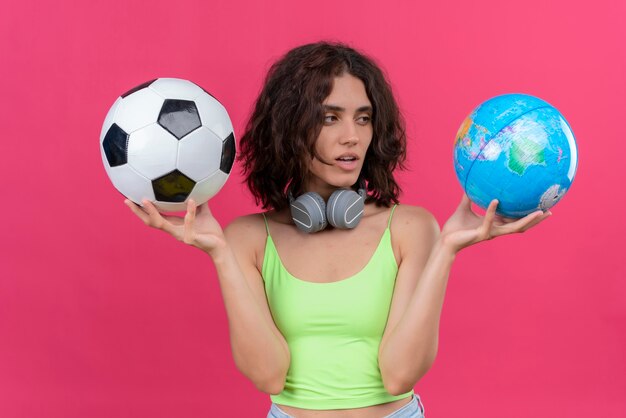 Una mujer joven y bonita con el pelo corto en la parte superior verde de la cosecha en auriculares con globo y balón de fútbol