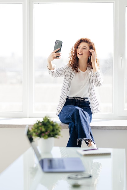 Una mujer joven y bonita con el pelo largo y rojo se toma una selfie en su teléfono inteligente mientras se sienta en el alféizar de la ventana Hermosa chica con un teléfono en un entorno doméstico Concepto de tecnología y redes sociales