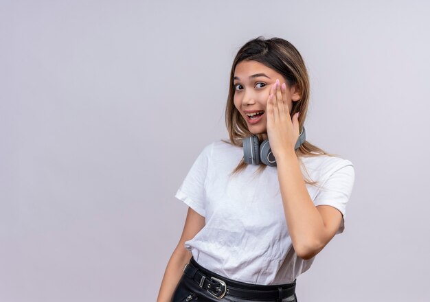 Una mujer joven y bonita positiva en camiseta blanca en auriculares manteniendo la mano en la cara mientras mira en una pared blanca