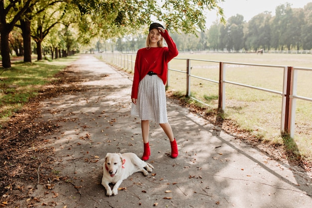 Mujer joven y bonita que parece feliz con su perro encantador al aire libre. Rubia alegre en jersey rojo y zapatos elegantes divirtiéndose en el parque de otoño.