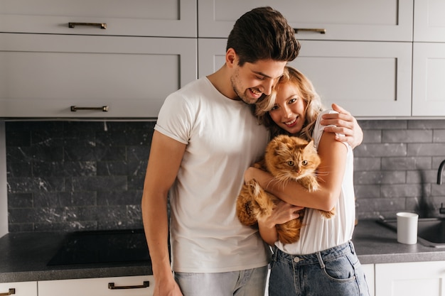 Mujer joven y bonita que sostiene el gato durante el retrato familiar. Lindo hombre morena abrazando a su esposa.