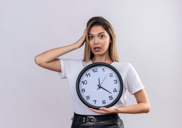 Una mujer joven y bonita sorprendida en camiseta blanca manteniendo la mano en la cabeza mientras sostiene el reloj de pared