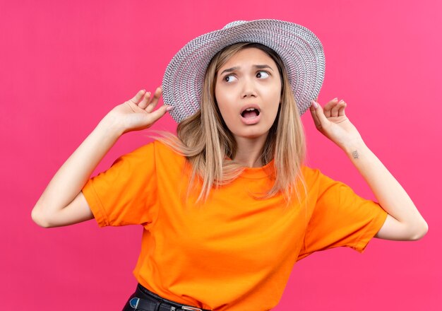 Una mujer joven y bonita sorprendida en una camiseta naranja con sombrero para el sol manteniendo la mano en su sombrero mientras mira de lado en una pared rosa