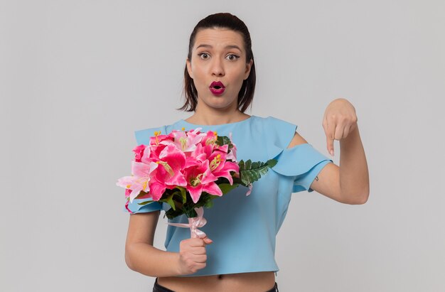 Mujer joven y bonita sorprendida sosteniendo un ramo de flores y apuntando hacia abajo