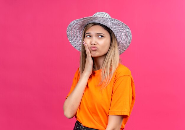 Una mujer joven y bonita triste con una camiseta naranja con sombrero para el sol manteniendo la mano sobre los dientes mientras tiene dolor en una pared rosa