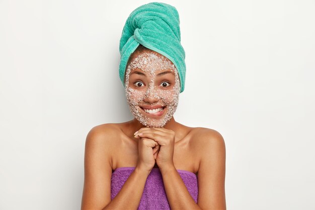 Una mujer joven y bonita se ve alegre, tiene una sonrisa agradable con dientes, muestra dientes perfectos y uniformes, mantiene las manos debajo de la barbilla, disfruta del tratamiento facial, aplica una máscara de sal marina útil para lucir saludable