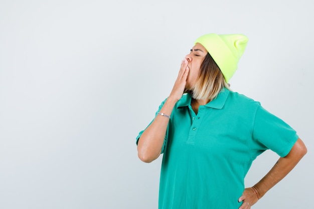 Mujer joven bostezando mientras mantiene la mano en la cadera en una camiseta de polo, gorro y con aspecto soñoliento. vista frontal.