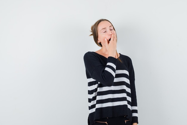 Mujer joven bostezando en prendas de punto a rayas y pantalón negro y con aspecto cansado