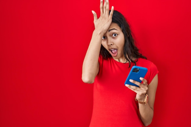 Foto gratuita mujer joven brasileña usando teléfono inteligente sobre fondo rojo sorprendida con la mano en la cabeza por error recuerda error olvidó concepto de mala memoria