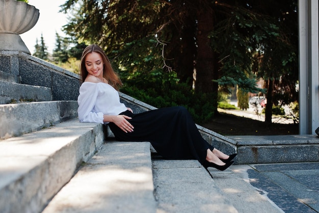 Mujer joven de buen aspecto con blusa blanca, pantalones negros anchos y tacones altos clásicos negros, sentada en las escaleras y posando