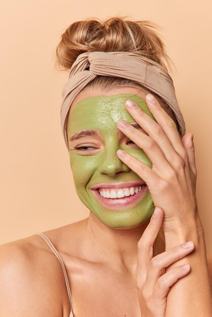 Una mujer joven de buen aspecto optimista cubre la cara con una sonrisa de mano y aplica ampliamente una máscara verde natural en la cara que se siente feliz de usar una diadema que se encuentra medio desnuda en el interior. Concepto de belleza y bienestar spa