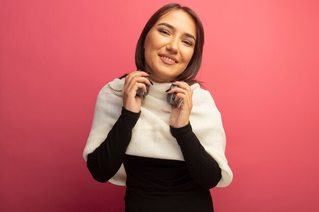 Mujer joven con bufanda blanca y auriculares alrededor de su cuello mirando a la cámara sonriendo alegremente