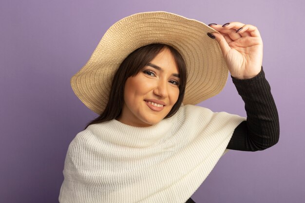 Mujer joven con bufanda blanca y sombrero de verano mirando al frente feliz y positivo tocando su sombrero de pie sobre la pared púrpura