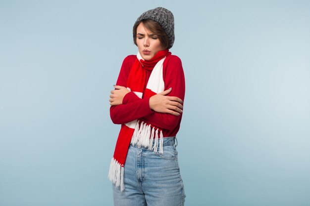 Mujer joven con cabello corto oscuro en suéter rojo y gorro de punto temblando de frío sobre fondo azul aislado