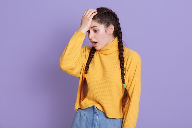 mujer joven con cabello largo y oscuro y coletas de pie con la boca abierta, manteniendo la mano en la frente