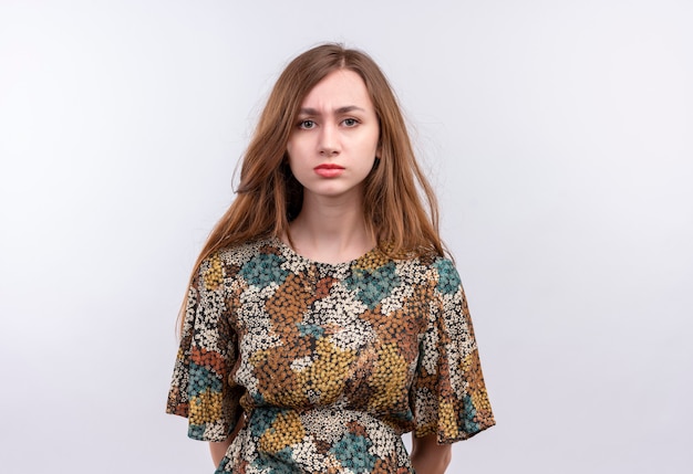 Mujer joven con cabello largo vistiendo coloridos vestidos muy mirando con expresión triste en la cara de pie sobre la pared blanca