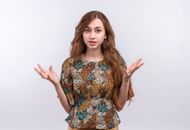 Mujer joven con cabello largo vistiendo coloridos vestidos de pie con los brazos levantados mirando confundido sobre la pared blanca