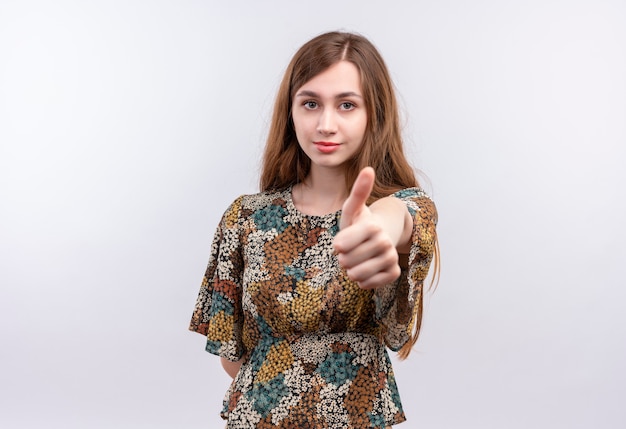 Mujer joven con cabello largo vistiendo coloridos vestidos sonriendo seguros mostrando Thumbs up de pie sobre la pared blanca