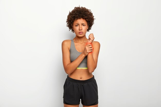 Mujer joven con cabello liso, toca la muñeca lesionada, sufre de sensaciones desagradables, vestida con ropa casual