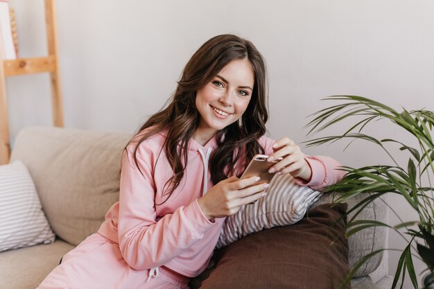 Mujer joven con cabello oscuro vestida con una sudadera con capucha rosa, está sentada en su sofá suave y viendo nuevas fotos en su teléfono inteligente