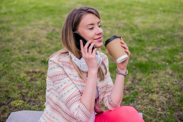 Mujer joven con café para ir en la mano usando un teléfono móvil y auriculares en el parque. Joven, elegante, hermoso, niña, Escuchar, Música, móvil, teléfono, auriculares, gozar, sonriente, feliz, fresco, accesorios, vendimia, estilo, divirtiéndose, riendo