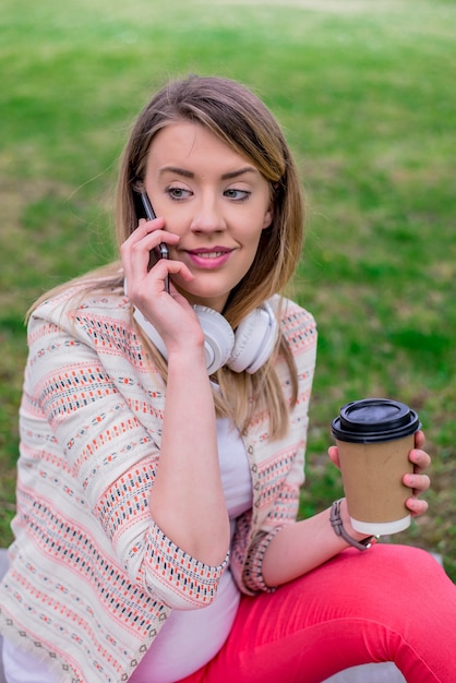 Mujer joven con café para ir en la mano usando un teléfono móvil y auriculares en el parque. Joven, elegante, hermoso, niña, Escuchar, Música, móvil, teléfono, auriculares, gozar, sonriente, feliz, fresco, accesorios, vendimia, estilo, divirtiéndose, riendo