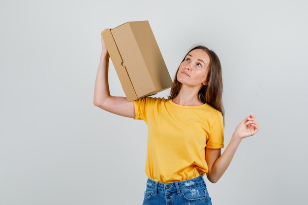 Foto gratuita mujer joven con caja de cartón y mirando hacia arriba en camiseta, vista frontal de pantalones cortos.