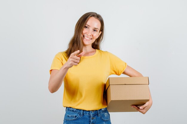 Mujer joven con caja de cartón con signo de dedo en camiseta, pantalones cortos y mirando feliz