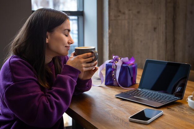 Mujer joven con caja de regalo y café delante de la computadora portátil