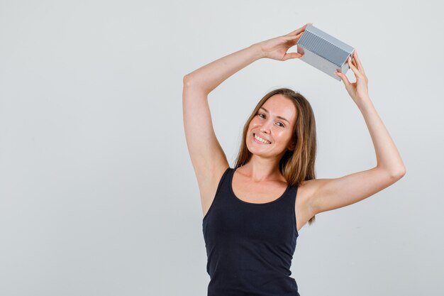 Mujer joven con caja de regalo sobre la cabeza en camiseta y mirando alegre. vista frontal.