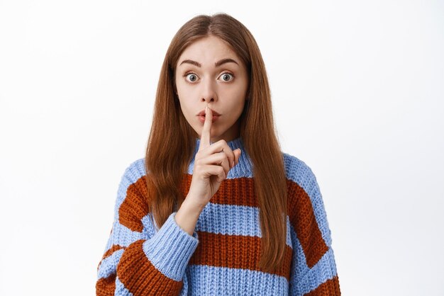 Mujer joven callando con el dedo presionado en los labios pidiendo mantener en secreto preparar sorpresa haciendo el signo de shh hush de pie sobre fondo blanco.