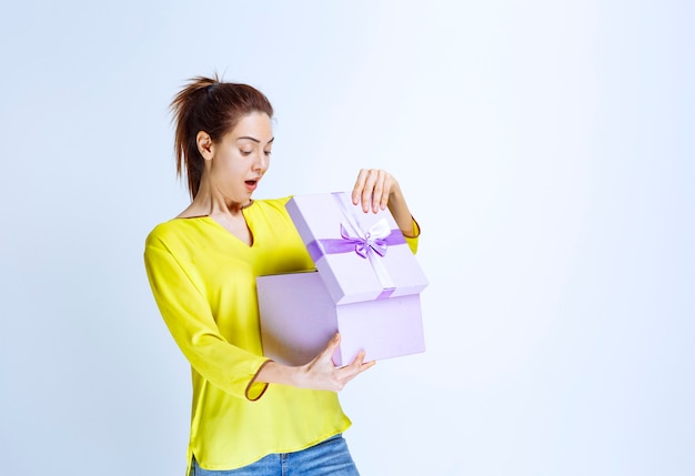 Mujer joven en camisa amarilla abriendo una caja de regalo violeta con sorpresa