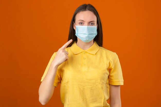 Mujer joven con camisa amarilla en máscara médica protectora apuntando con el dedo en su máscara con cara seria mirando a la cámara de pie sobre fondo naranja