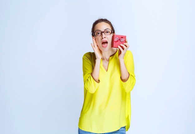 Mujer joven con camisa amarilla mostrando su caja de regalo roja y mirando sorprendido