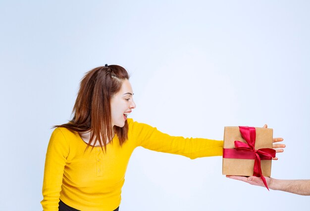 Mujer joven con camisa amarilla se ofrece una caja de regalo de cartón con cinta roja y mano anhelante para tomarla