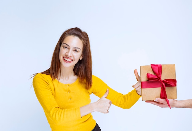 A una mujer joven con camisa amarilla se le ofrece una caja de regalo de cartón con cinta roja y muestra un signo de mano positivo