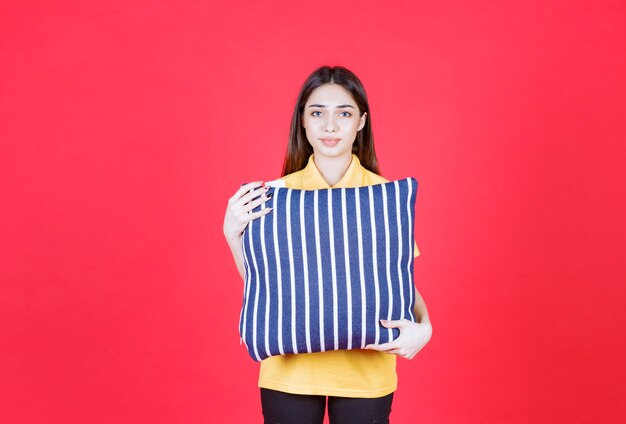 Mujer joven en camisa amarilla sosteniendo una almohada azul con rayas blancas