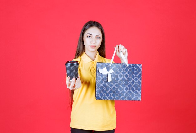 Mujer joven en camisa amarilla sosteniendo una bolsa azul y una taza de bebida desechable negra