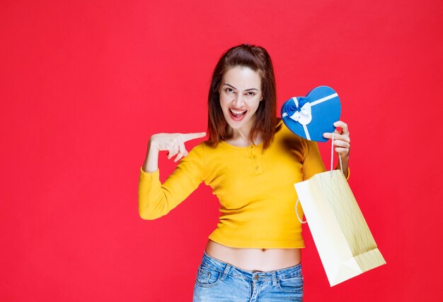 Mujer joven con camisa amarilla sosteniendo una bolsa de cartón, tomando una caja de regalo azul y sintiéndose sorprendida