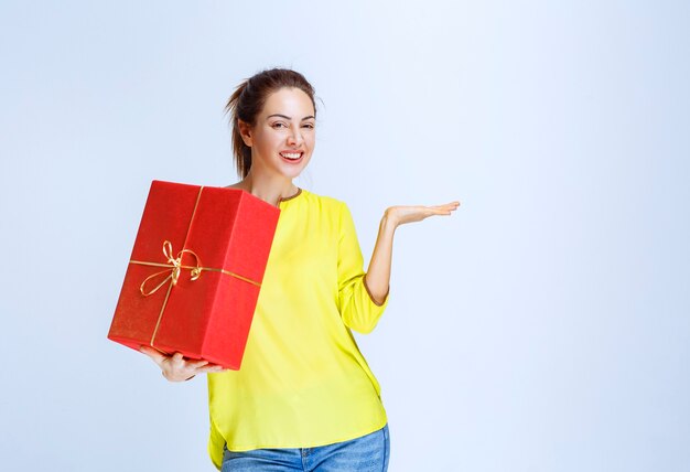 Mujer joven en camisa amarilla sosteniendo una caja de regalo roja y apuntando a ella