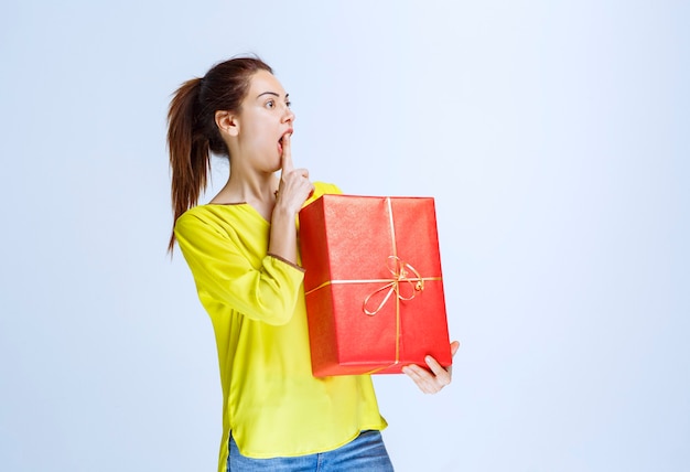 Mujer joven en camisa amarilla sosteniendo una caja de regalo roja y pensando o dudando