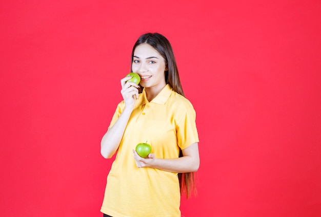 Mujer joven en camisa amarilla sosteniendo una manzana verde y tomando un bocado
