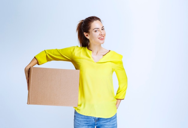 Foto gratuita mujer joven en camisa amarilla sosteniendo su caja de carga de cartón y sintiéndose feliz