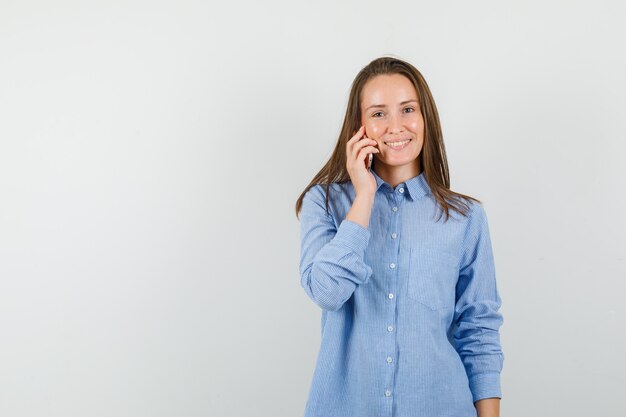 Mujer joven con camisa azul hablando por teléfono móvil y luciendo optimista