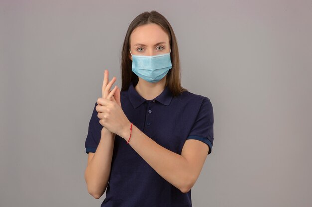 Mujer joven en camisa azul marino y máscara protectora médica mostrando manos limpias mirando a la cámara de pie sobre fondo gris claro