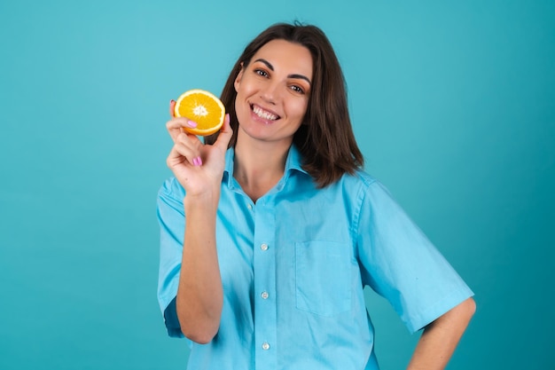 Mujer joven con una camisa azul en la pared sostiene una naranja, posa alegremente, de buen humor, se ríe, sonríe