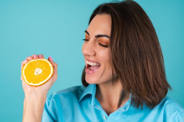 Mujer joven con una camisa azul en la pared sostiene una naranja, posa alegremente, de buen humor, se ríe, sonríe