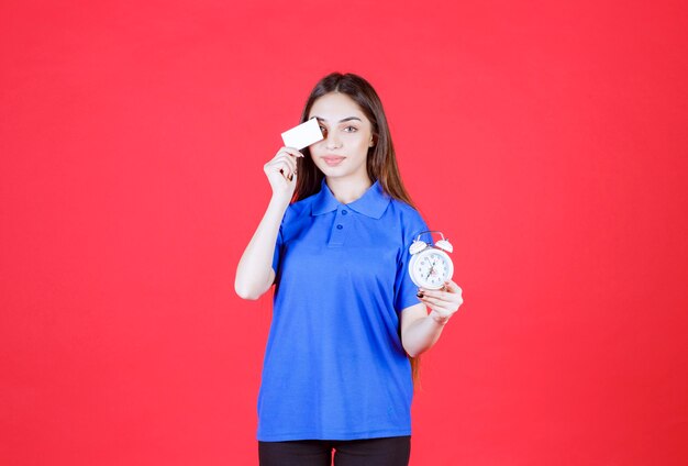 Mujer joven con camisa azul sosteniendo un reloj despertador y presentando su tarjeta de visita