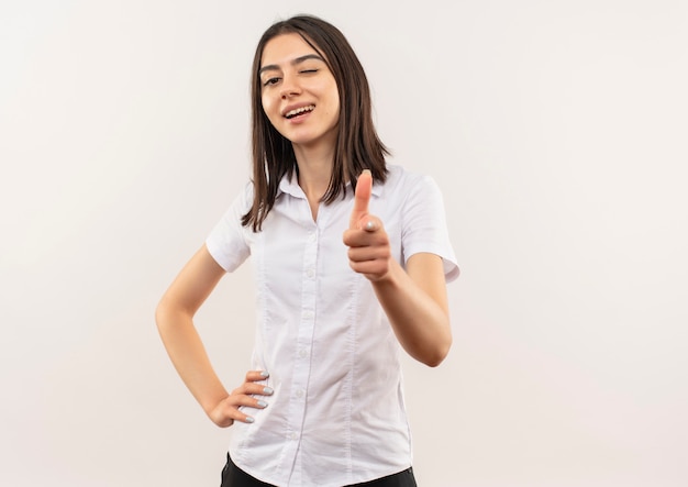 Mujer joven con camisa blanca apuntando con el dedo hacia el frente sonriendo y guiñando un ojo de pie sobre la pared blanca