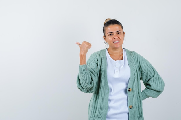 Mujer joven con camisa blanca y chaqueta de punto verde menta apuntando hacia atrás mientras sostiene la mano detrás de la cintura y parece feliz